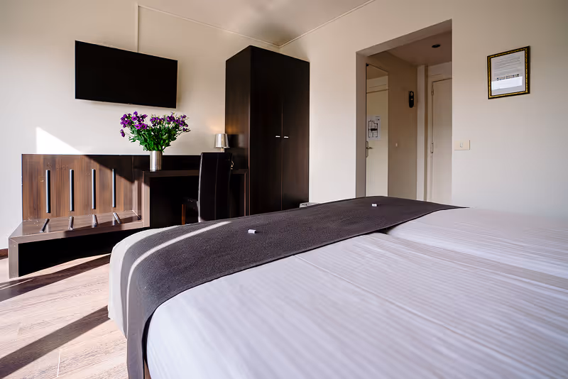 Hotel room with two beds covered in white linens and dark runners, a wall-mounted flat-screen TV, a desk with a chair, a vase of purple flowers, and a wardrobe.