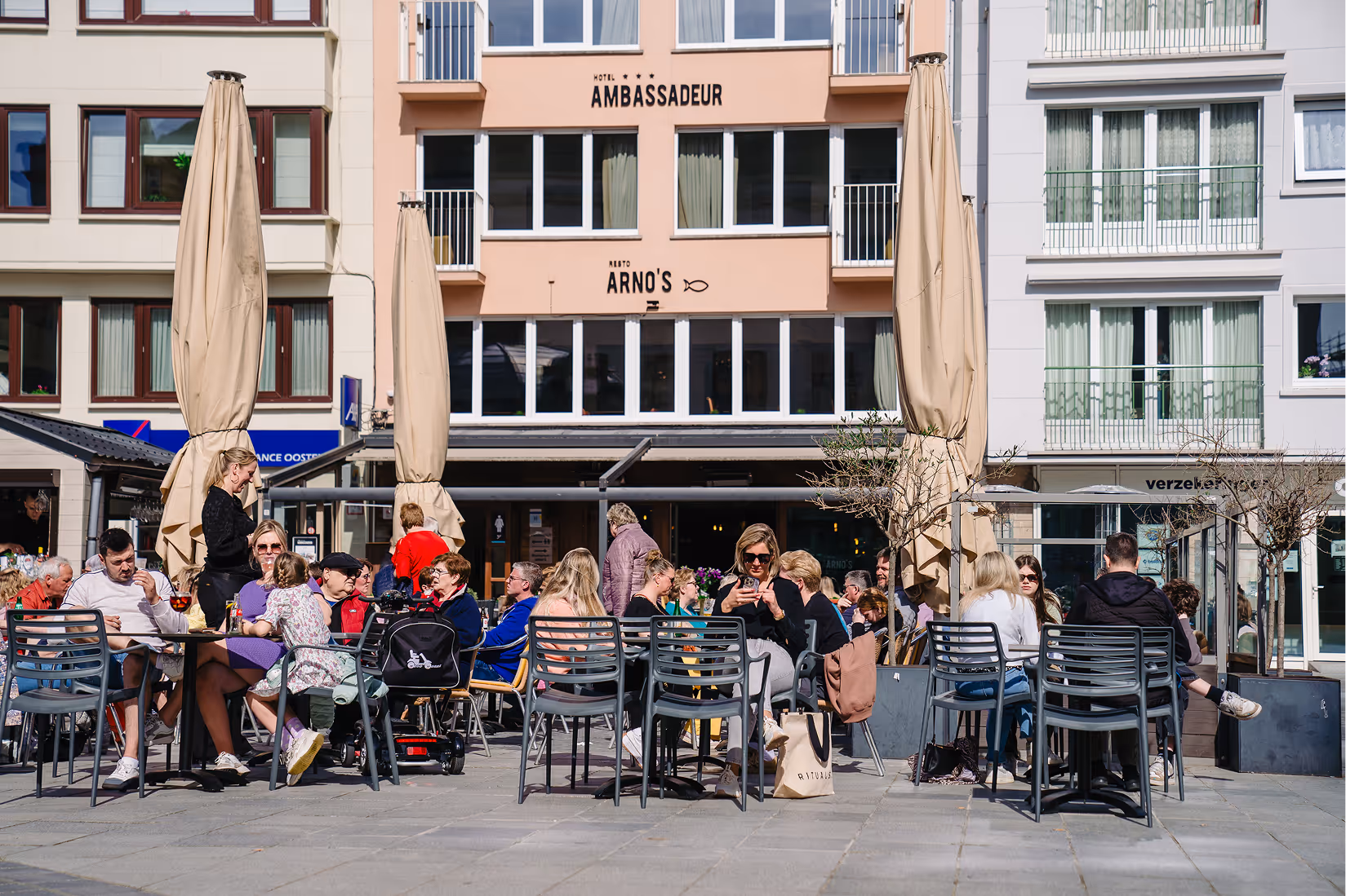Outdoor café seating area with people dining and socializing on a sunny day in front of Hotel Ambassadeur and Resto Arno's.