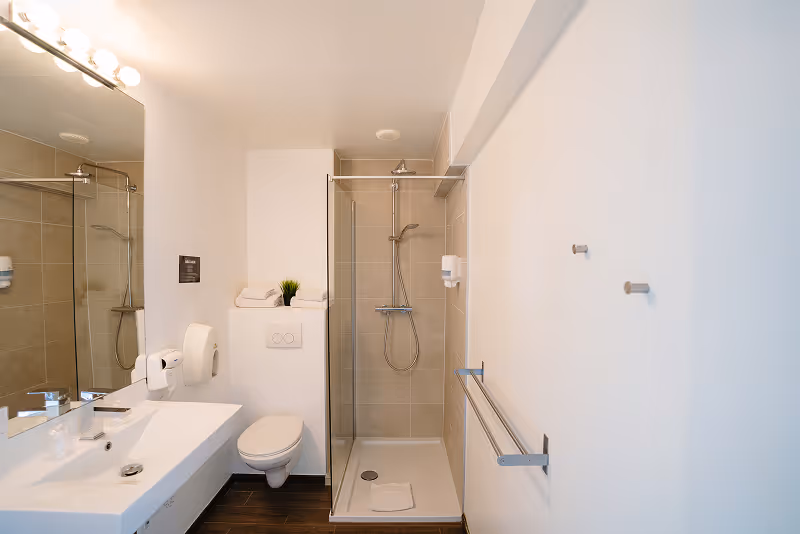 Modern bathroom with a white sink, mounted toilet, glass-enclosed shower, white walls, and wooden floor.